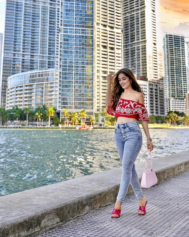Roopsi Bansal in a red embroidered off-shoulder ruffle crop top and ripped skinny jeans from Express, holding a pink handbag, standing along the water at Bayfront with the Miami skyline behind her