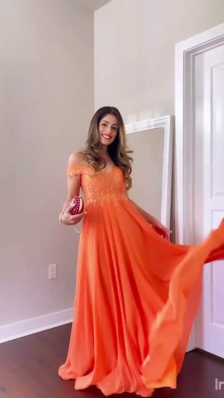 Roopsi Bansal in an orange off-shoulder chiffon gown with a lace bodice, twirling in front of a mirror, holding a beaded clutch