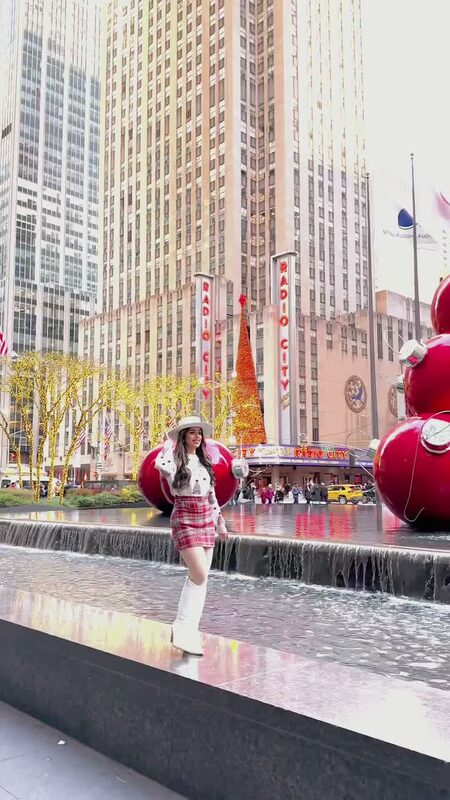 Roopsi Bansal in a white sweater and red plaid mini skirt from Chicwish beside giant red ornament decorations near Radio City Music Hall, NYC