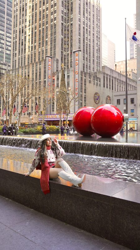 Roopsi Bansal in a cream Christmas tree pattern sweater set sitting by a fountain beside two giant red ornaments on 6th Avenue in New York