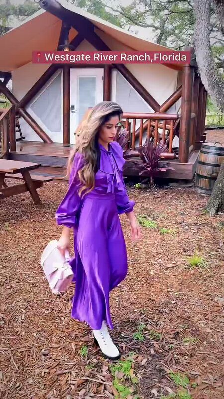 Roopsi Bansal in a purple satin bow-neck dress from SHEIN and white lace-up boots, carrying a lilac handbag, standing outside a canvas glamping tent at Westgate River Ranch Resort