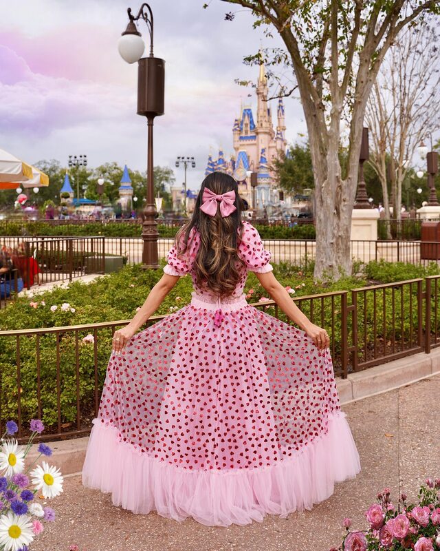 Roopsi Bansal in a pink heart-print puffed-sleeve tulle gown with a pink bow, back to the camera, with Cinderella Castle visible in the distance at Walt Disney World
