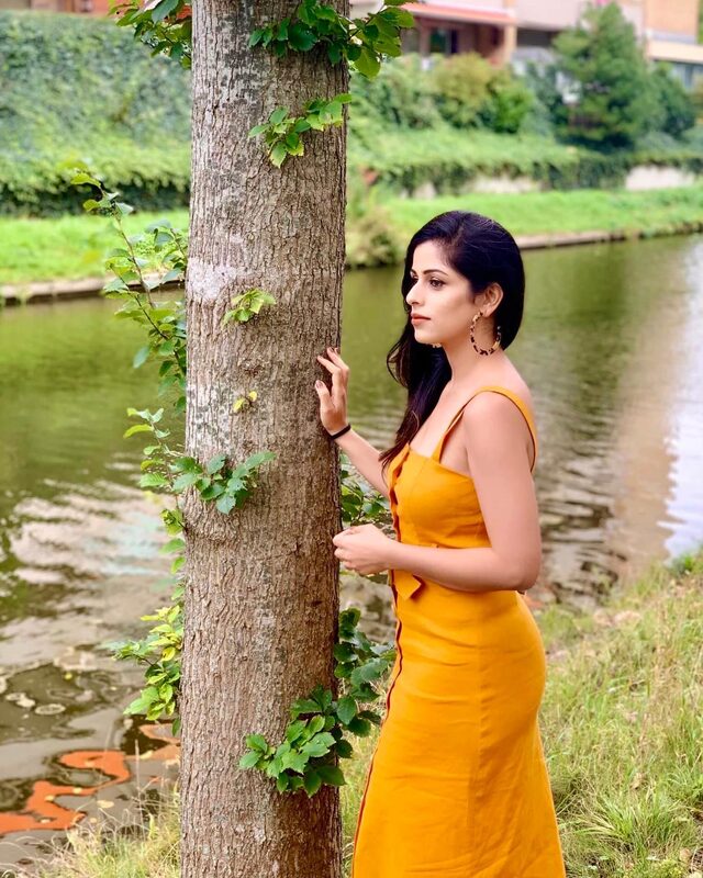 Standing beside a tree along a green canal in a mustard-yellow slip dress and tortoiseshell hoop earrings, one hand resting on the bark