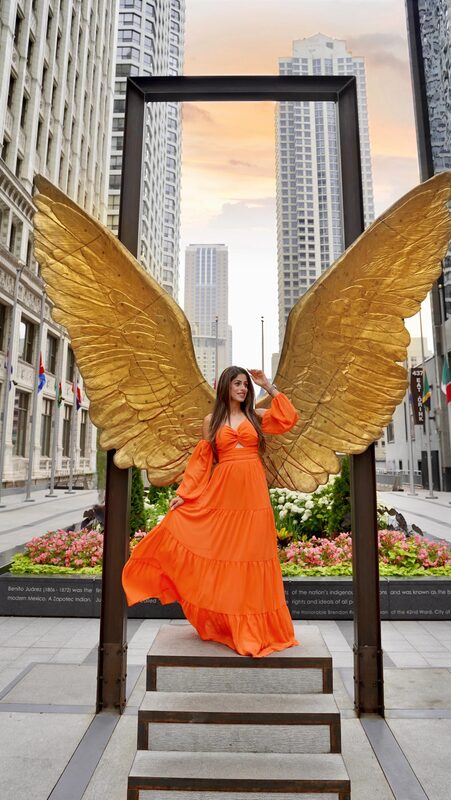 Roopsi Bansal in an orange tiered dress standing in front of a giant golden angel-wing sculpture on Mag Mile, Chicago skyline glowing at sunset behind her