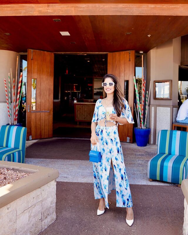 Roopsi Bansal in a blue tie-dye cutout maxi dress, holding a cocktail, standing in front of the wooden entrance doors of S3 Sun Surf Sand restaurant
