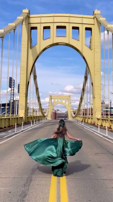 Roopsi Bansal from behind in a flowing emerald satin gown, standing under the yellow arch of the Roberto Clemente Bridge in Pittsburgh