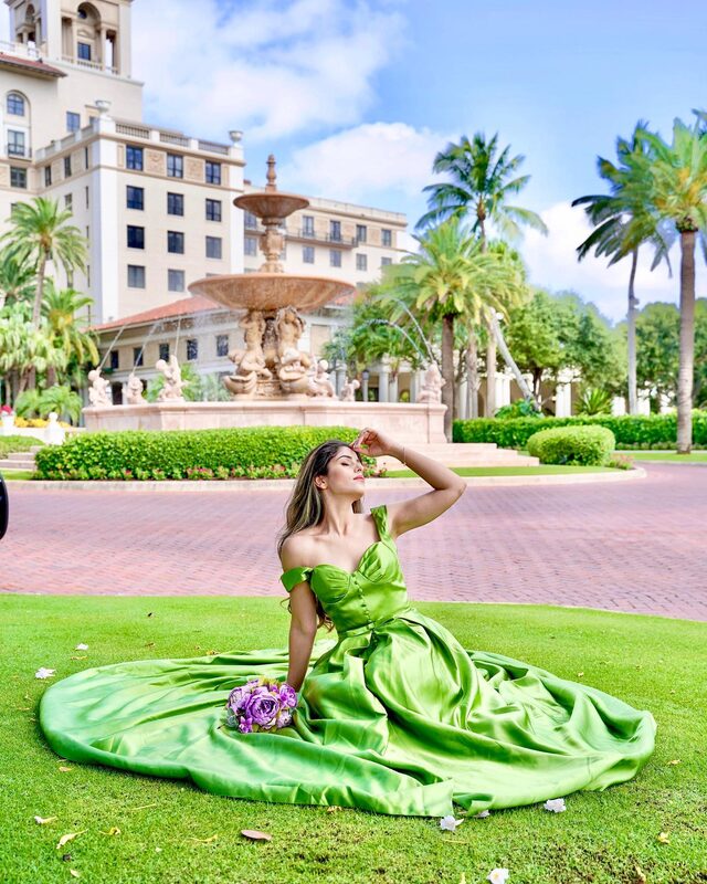 Roopsi Bansal in a vivid green off-shoulder ball gown, seated on the lawn in front of the grand fountain and Mediterranean-style building at The Breakers Palm Beach