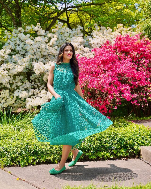 Roopsi Bansal in a twirling emerald green lace dress in front of white and hot pink azalea bushes in full bloom