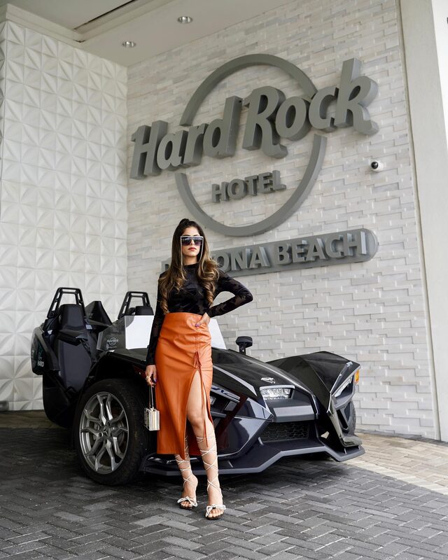 Roopsi Bansal in a black mesh top and orange faux-leather midi skirt with a front slit, standing beside a black three-wheeled sports car under the Hard Rock Hotel Daytona Beach sign