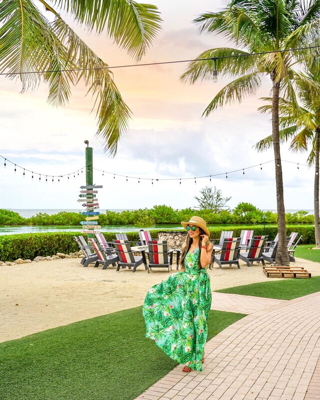 Roopsi Bansal in a green tropical print maxi dress and straw hat, walking a garden path lined with palm trees and string lights at Hawks Cay Resort