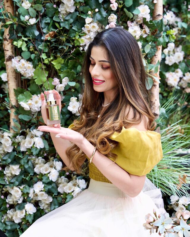 Roopsi Bansal in a mustard off-shoulder top holding a bottle of Hairssential hair oil in front of a wall of white roses
