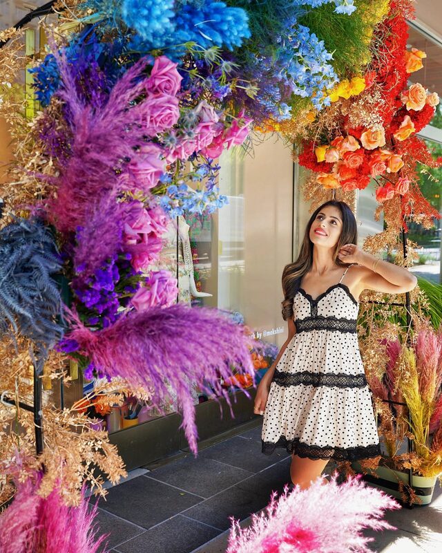 Roopsi Bansal in a black and white polka dot lace-trim trapeze dress from Express, standing beneath a floral archway of pink, blue, and purple flowers in Buckhead, Georgia