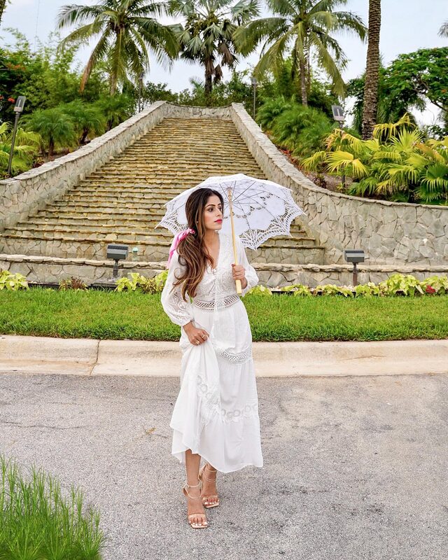 Roopsi Bansal in a white lace-up maxi dress with crochet trim, holding a white lace parasol, standing in front of a wide stone stairway lined with palm trees