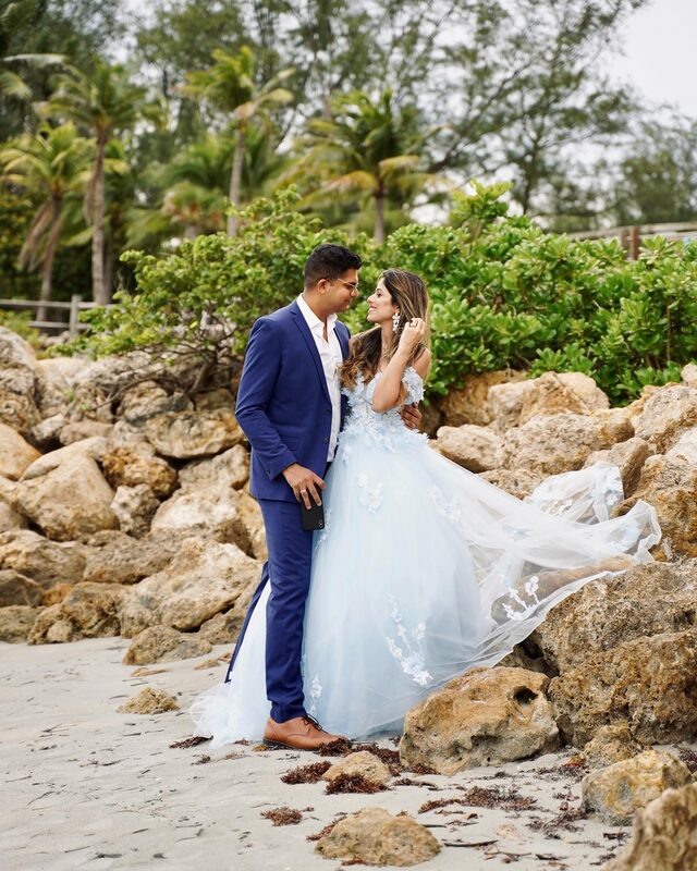Roopsi Bansal in a powder blue floral applique gown and her husband in a navy suit, standing face to face among coral rocks on a beach