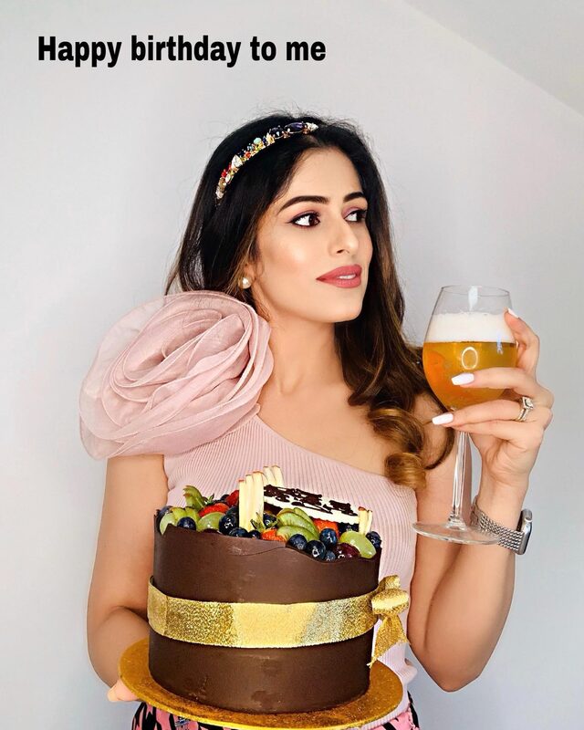 Holding a chocolate birthday cake topped with fresh fruit and wrapped in a gold ribbon, wearing a pink one-shoulder top with a large fabric rose and a jeweled headband, holding a glass of something bubbly