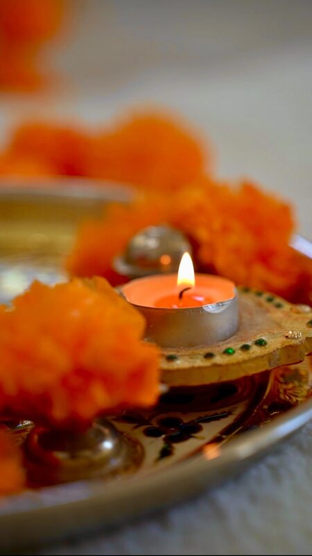 A close-up of a lit diya candle surrounded by orange marigold flowers on a decorated brass plate