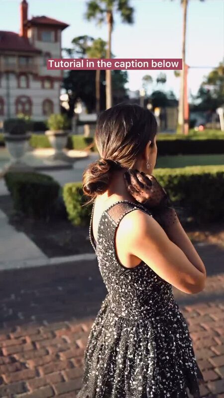 Roopsi Bansal in a black sequined dress and sheer polka-dot gloves, hand at her low bun, seen from behind in front of an ornate stone building