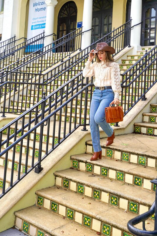 Roopsi Bansal in a cream lace blouse, brown cowboy hat, and jeans, descending green and yellow tiled steps outside a church at Rosemary Square, holding a rust-colored briefcase