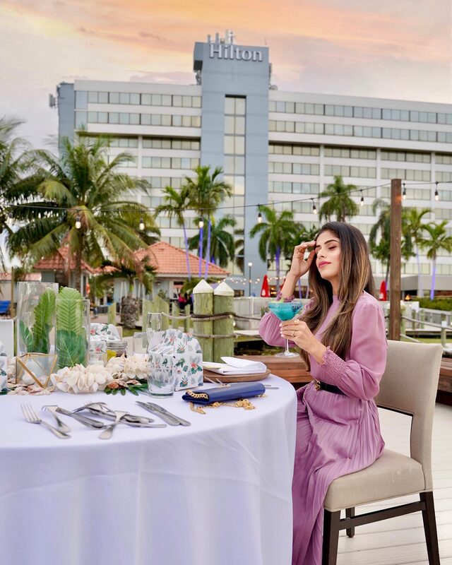 Roopsi Bansal in a mauve pleated long-sleeve dress, seated at a lakeside dinner table with a blue cocktail, the Hilton Palm Beach Airport tower lit up at sunset behind her