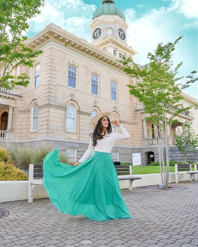 Roopsi Bansal in a white full-sleeve top, mint green chiffon maxi skirt, and straw boater hat, twirling in front of the clock tower of Athens City Hall, Georgia