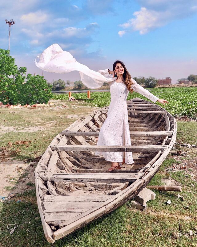 Roopsi Bansal in a white embroidered kurta, her white dupatta caught in the wind, standing in an old wooden boat beside a green field