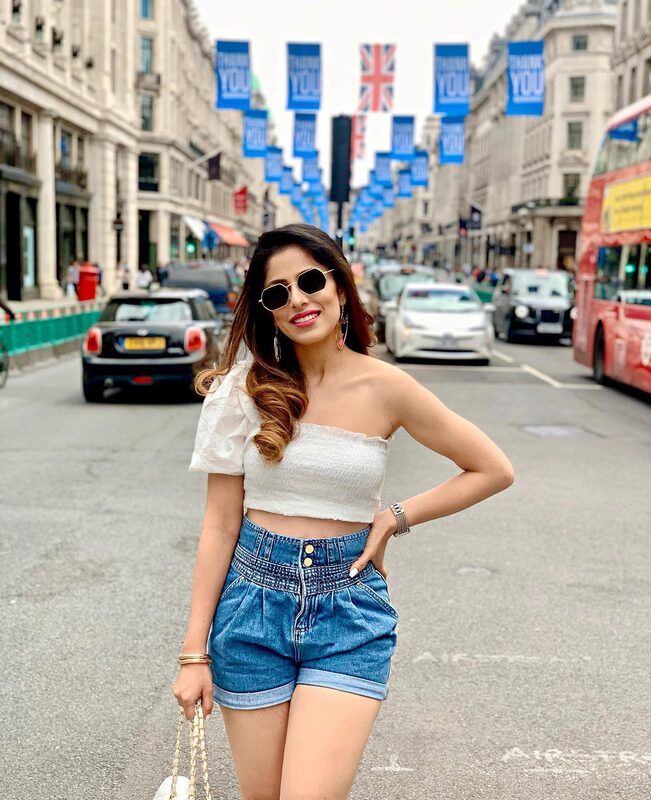 Roopsi Bansal in a white one-shoulder crop top and denim shorts, standing on Regent Street in London under rows of Union Jack banners