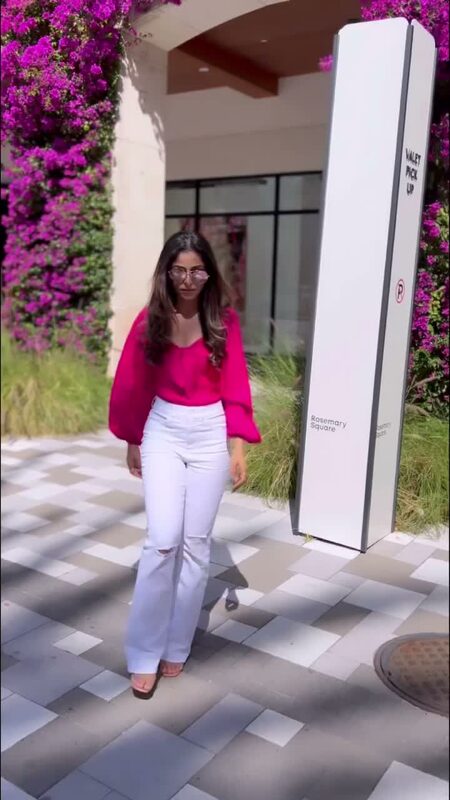 Roopsi Bansal in a hot pink puff-sleeve blouse and white ripped flare jeans from Express, wearing round sunglasses, walking past a wall of purple bougainvillea