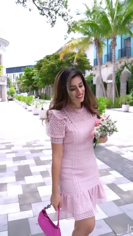 Roopsi Bansal in a pink lace dress carrying a hot pink handbag and a small bouquet, walking down a palm-lined plaza