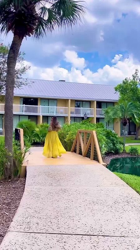 Roopsi Bansal in a golden yellow full ballgown skirt from Chicwish, walking across a wooden footbridge over a pond at a resort