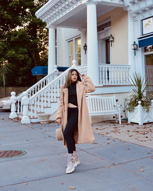 Roopsi Bansal in a beige teddy coat over a black crop set and snakeskin boots, standing in front of a white columned porch