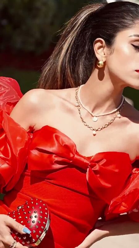 Roopsi Bansal in a red off-shoulder puff-sleeve dress with a bow at the bust, holding a red heart-shaped studded clutch, layered gold necklaces at her collarbone