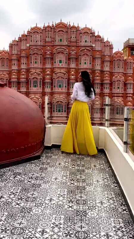 Roopsi Bansal in a yellow maxi skirt and white top standing on a rooftop in front of the Hawa Mahal's honeycomb pink sandstone facade in Jaipur