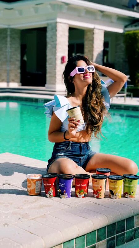 Roopsi Bansal in a white and light blue ruffled swimsuit, seated poolside behind a row of Revive Superfoods smoothie and soup cups