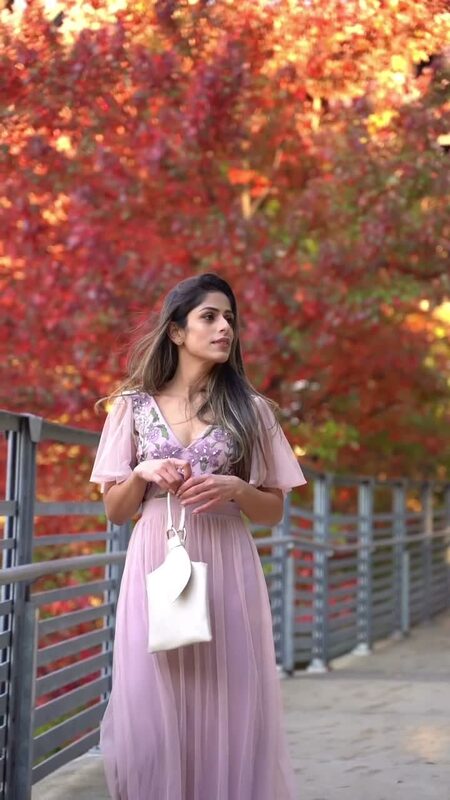 Roopsi Bansal in a blush floral-embroidered maxi dress on a footbridge surrounded by brilliant red and orange fall foliage