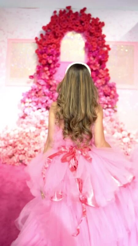 Roopsi Bansal from behind in a pink tiered tulle ball gown, standing beneath an arched doorway framed in red and pink roses