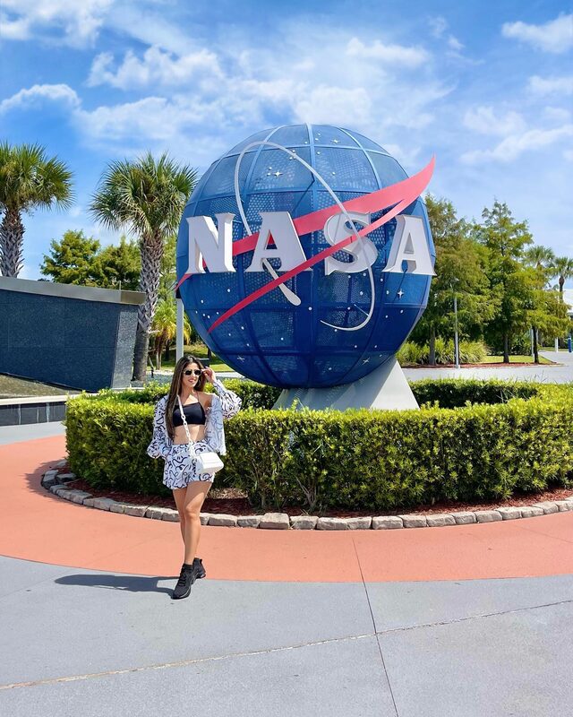 Roopsi Bansal in a black bralette with a white and blue graffiti-print jacket and matching shorts, standing in front of the large NASA meatball globe sculpture surrounded by palm trees