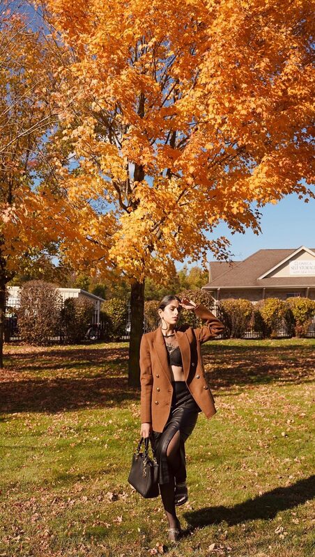 Roopsi Bansal in a camel double-breasted blazer, black lace bralette, and leather skirt standing under a bright orange fall tree