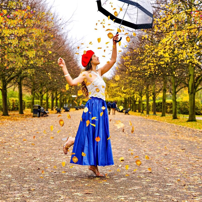 Roopsi Bansal in a red beret, white lace top and blue skirt, twirling with an umbrella as autumn leaves fall around her in a park