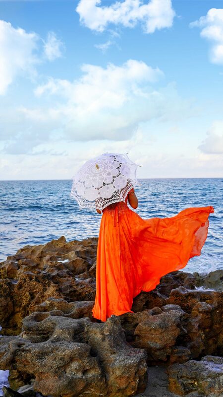 Roopsi Bansal from behind in a flowing orange gown, holding a white lace parasol, standing on the rocky shoreline at Coral Cove, Florida with the ocean ahead