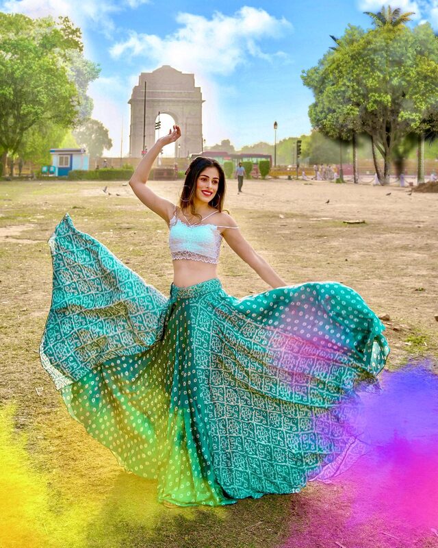 Roopsi Bansal twirling in a teal bandhani lehenga skirt and white lace crop top in front of India Gate in Delhi, with coloured Holi powder drifting in the air
