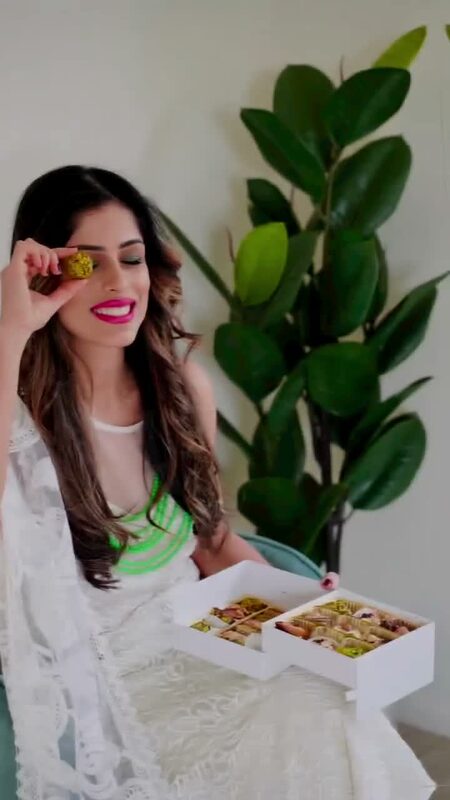 Roopsi Bansal holding up a pistachio sweet in front of a box of colorful Indian mithai from Layla's Delicacies