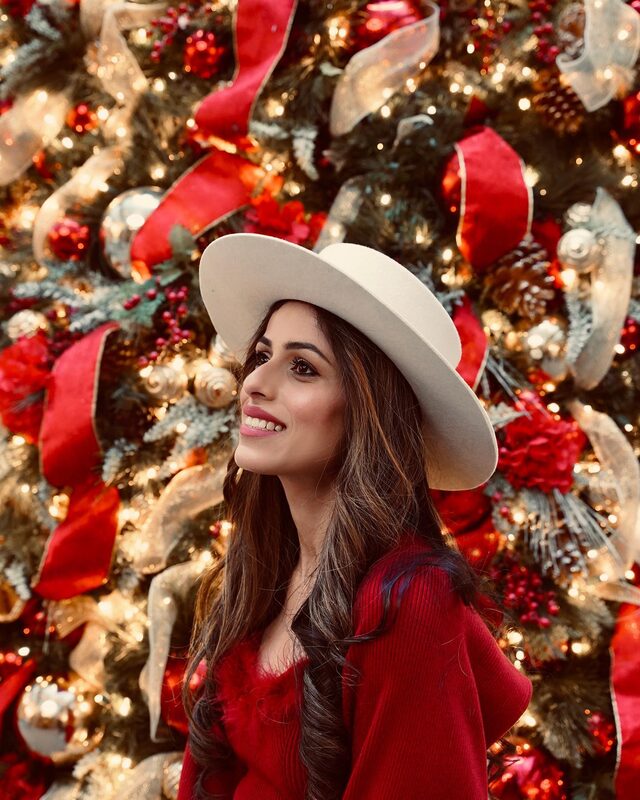 Roopsi Bansal in a cream wide-brim Gigi Pip hat and red sweater in front of a Christmas tree decorated in red and gold