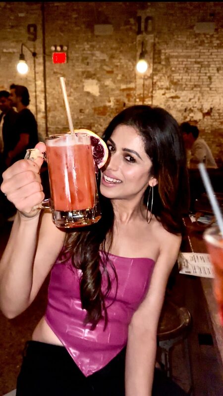 Roopsi Bansal holding up a large craft cocktail in a beer mug against a brick wall at Houston Hall