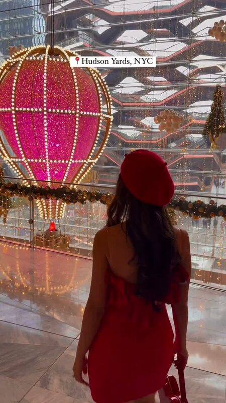 Roopsi Bansal in a red dress and red beret facing a light-wrapped pink hot air balloon installation inside the Vessel at Hudson Yards