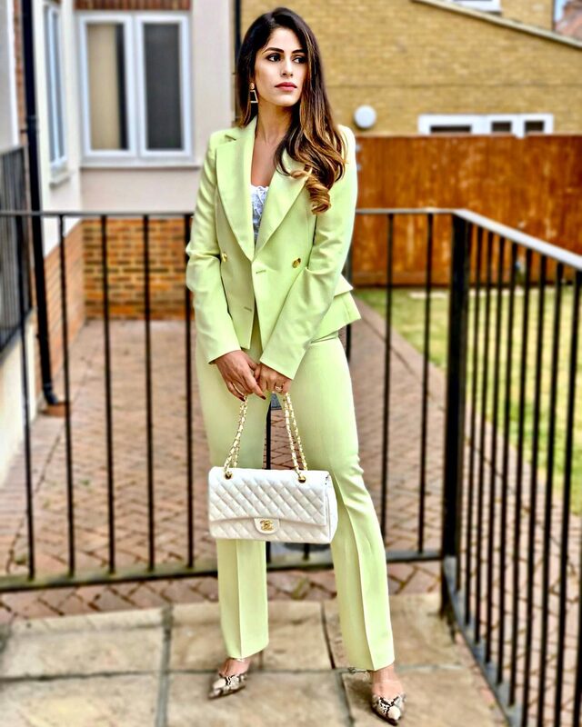 Standing by an iron railing wearing the same lime-green double-breasted blazer and trousers, carrying a white quilted Chanel flap bag