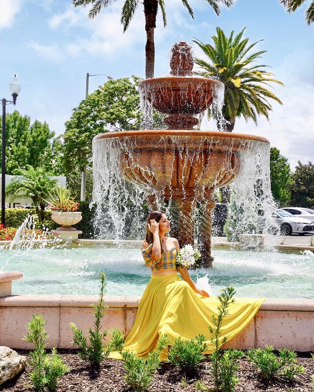 Roopsi Bansal in a teal lace-trimmed midi dress, holding a white lace parasol, standing under a wooden archway covered in purple bougainvillea in Orlando