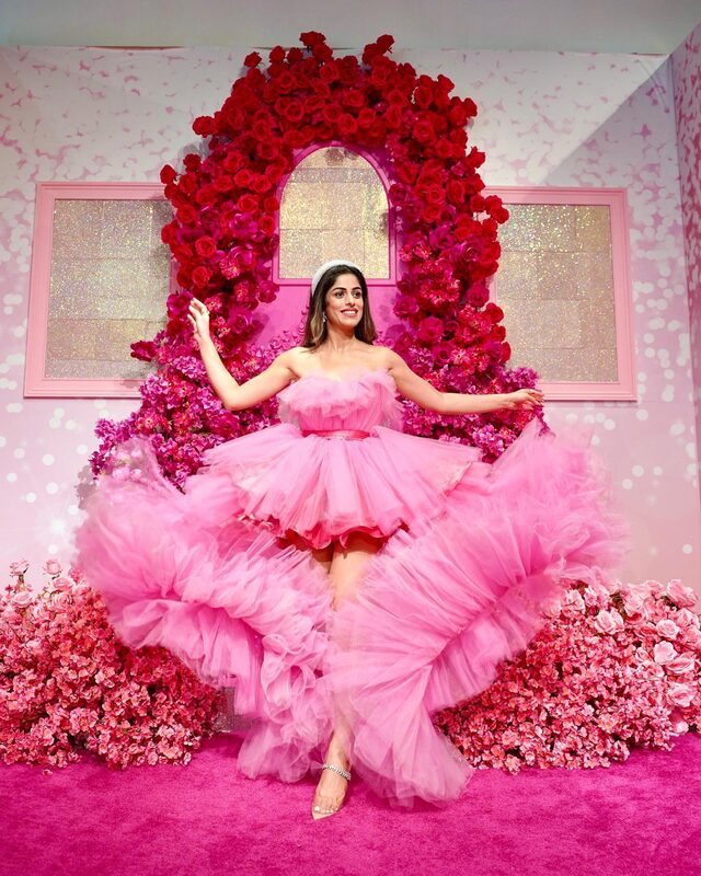 Roopsi Bansal in a strapless pink tulle ball gown with arms outstretched, standing in front of a red rose archway with a glittering arched window at a floral installation