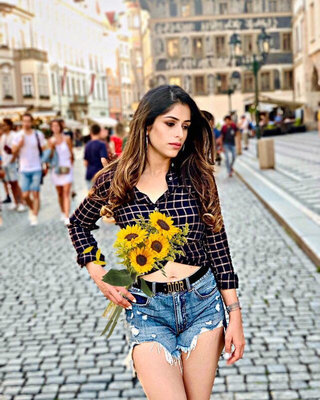Roopsi Bansal holding a bunch of sunflowers in a navy plaid shirt and denim shorts, standing in a cobblestone square in Prague, Czech Republic