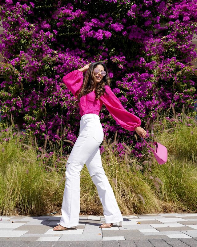Roopsi Bansal in a hot pink long-sleeve satin jacquard bodysuit and white high-waisted ripped bootcut jeans from Express, carrying a hot pink handbag and wearing mirrored sunglasses, in front of a wall of purple bougainvillea in West Palm Beach