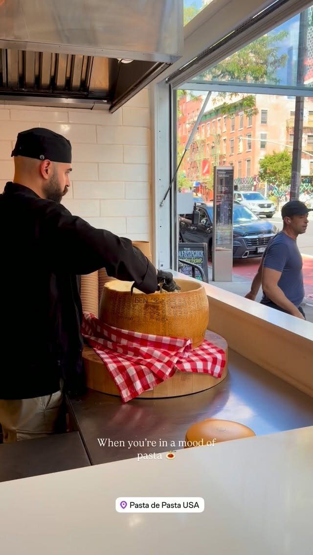 A chef shaving parmesan from a giant wheel at the window counter of Pasta de Pasta USA, with a New York City street visible through the glass